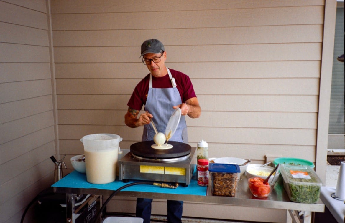 Man in glasses and an apron pours batter onto a round griddle to make a crepe, with bowls of toppings on a table beside him.
