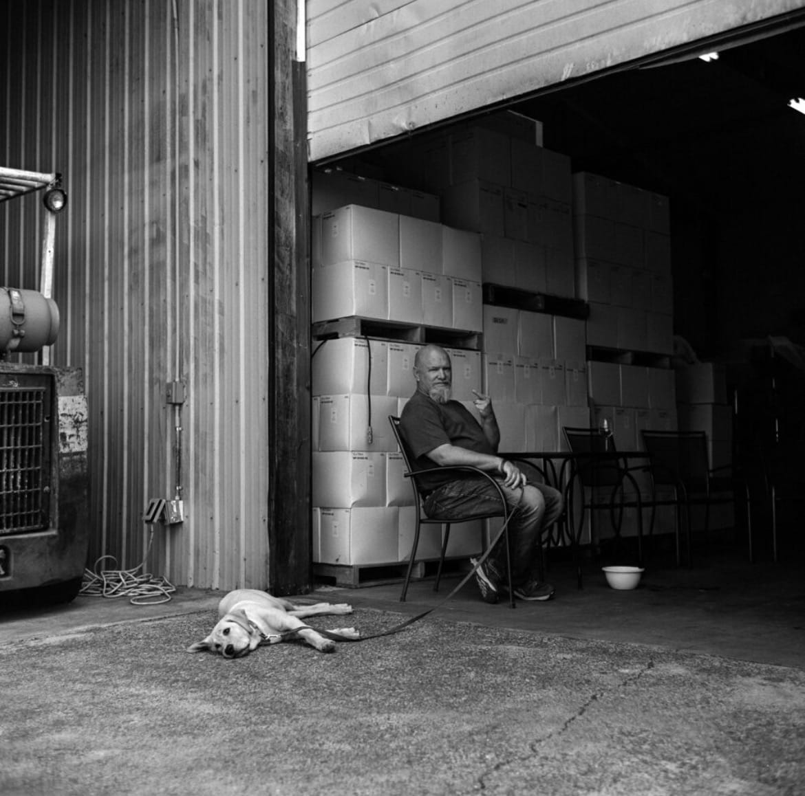 Black-and-white photo of a bearded man pointing at the camera from a chair in a warehouse doorway, a dog lying on a leash nearby.