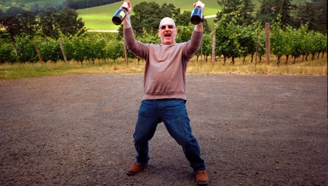 Man in sunglasses raises two wine bottles overhead on a gravel road, cheering in front of vineyard rows and forested hills.