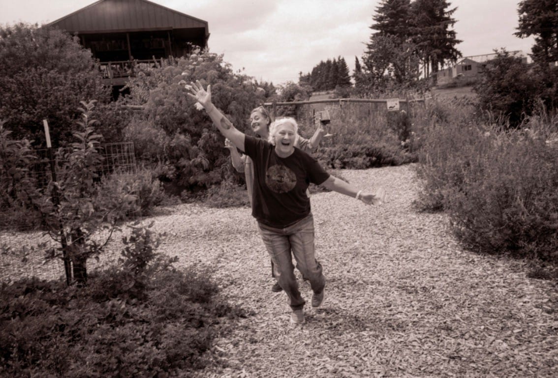 Sepia-toned photo of two laughing women with wine glasses running down a gravel garden path, arms outstretched, with a barn behind them.
