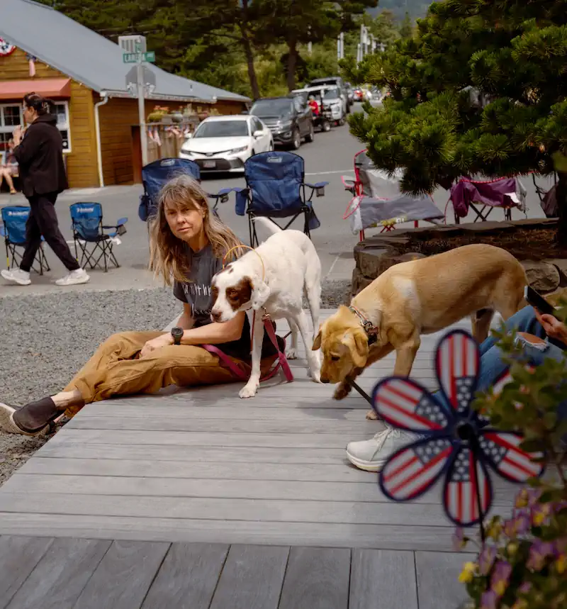 Woman sits on a wooden boardwalk with two dogs on leashes beside her, on a small-town street lined with camp chairs and parked cars.