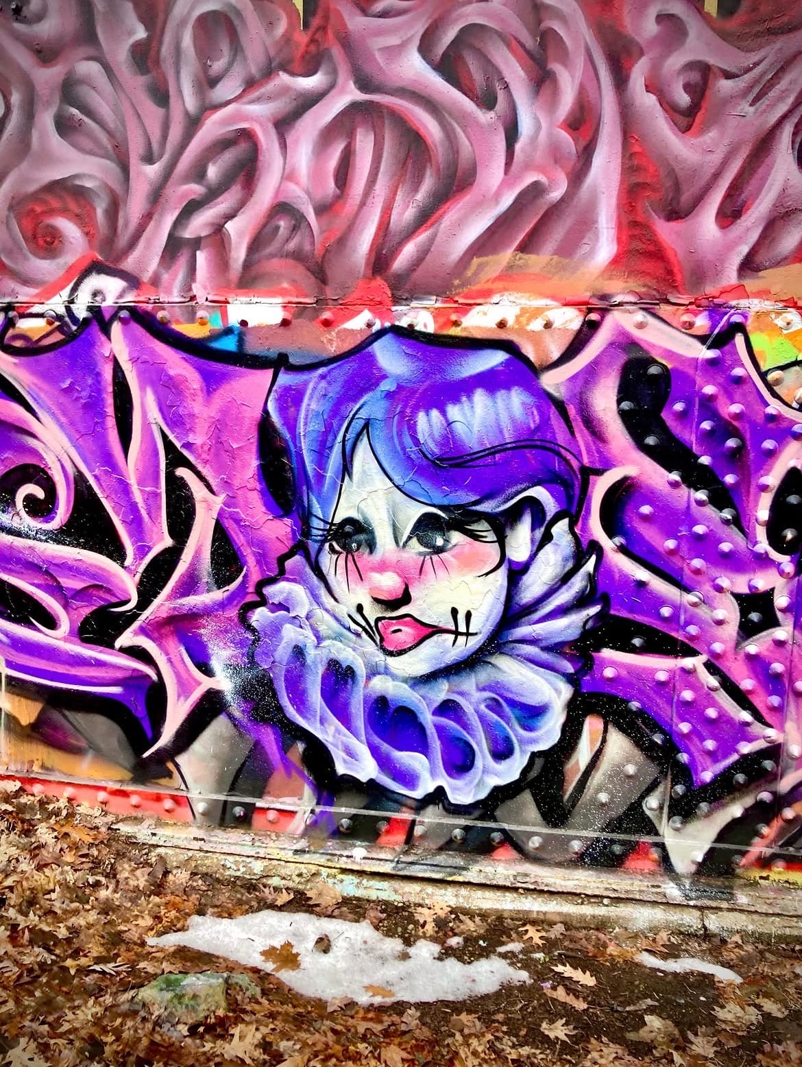 Spray-painted mural on riveted metal of a purple-haired jester-clown face with tears, framed by pink swirling patterns.