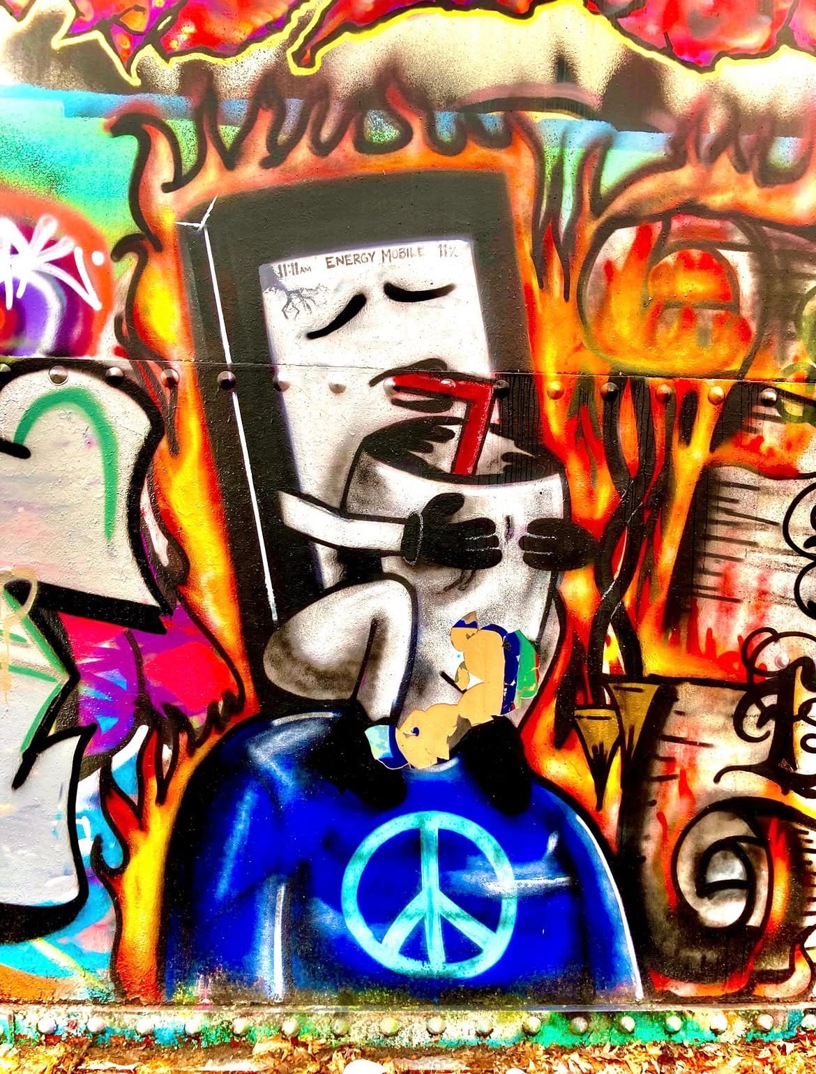 Spray-painted mural of a smartphone character labeled "Energy Mobile" drinking from a straw, engulfed in orange flames, wearing a blue peace-sign jacket.