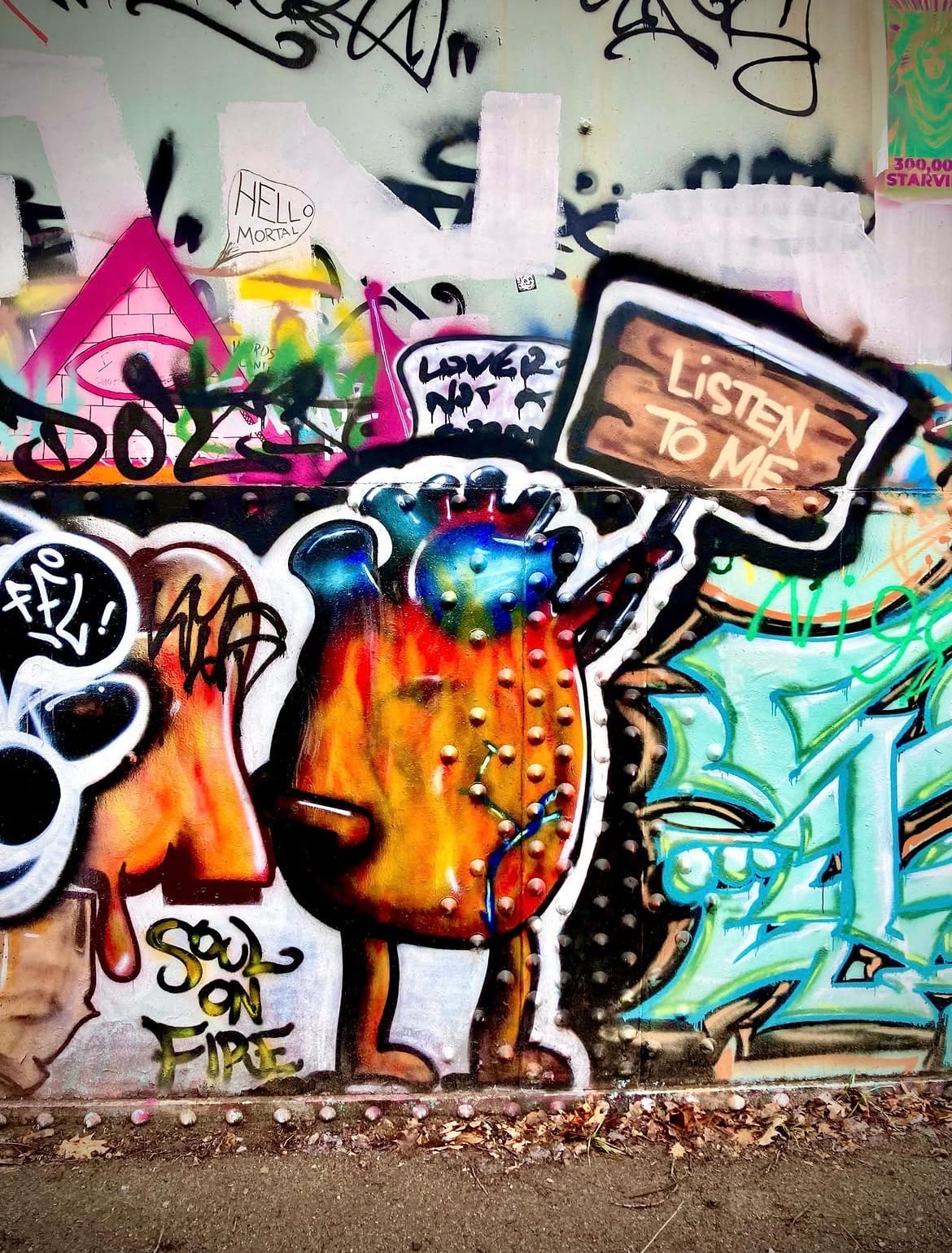 Spray-painted mural of a flame-bodied creature holding a sign reading "Listen to Me," beside graffiti text "Soul on Fire" and "Hello Mortal."