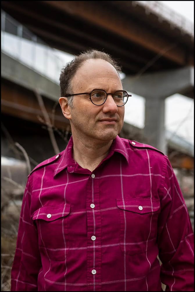 Photograph of a man in a magenta plaid shirt and round glasses looking off to the side, standing beneath a concrete highway overpass.