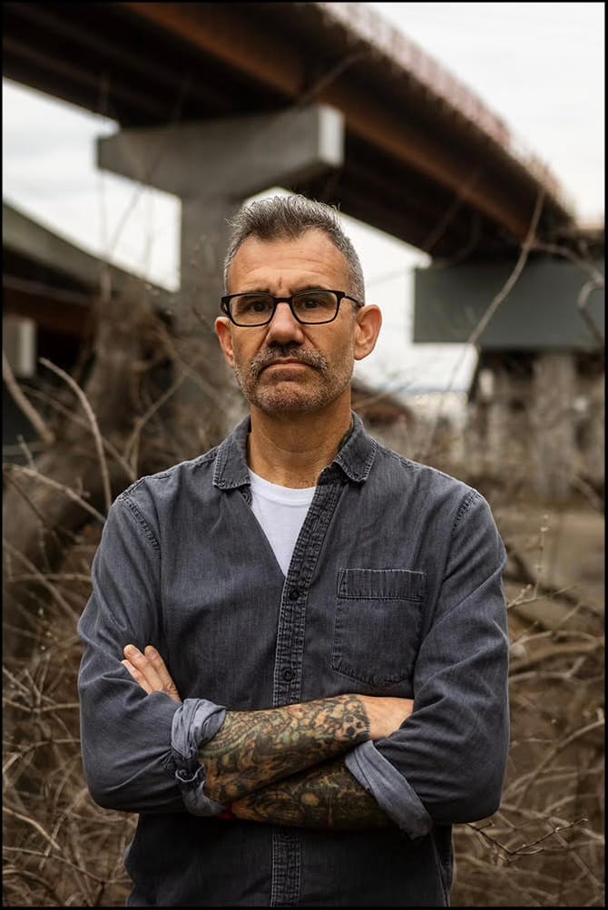 Photograph of a gray-haired, tattooed man in a denim shirt standing with arms crossed beneath a highway overpass.