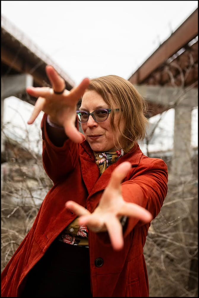 Photograph of a woman in a rust-colored coat reaching both hands toward the camera, standing beneath a highway overpass.