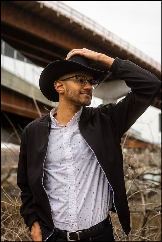 Photograph of a man in a black cowboy hat and jacket touching his brim, glancing sideways beneath a highway overpass.