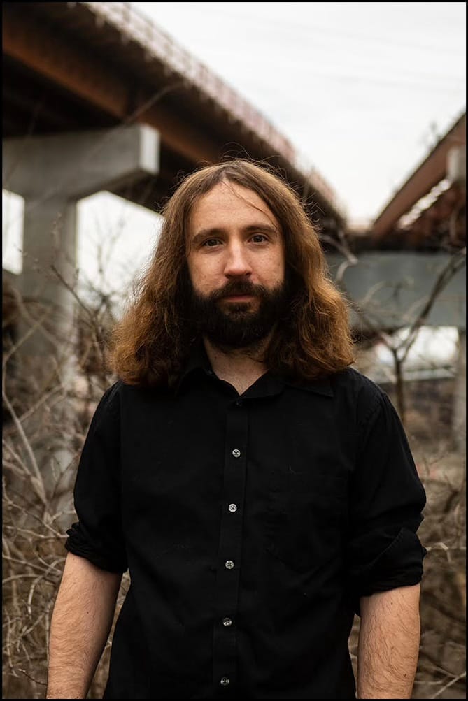 Photograph of a long-haired, bearded man in a black button-up shirt standing before dry brush beneath a highway overpass.