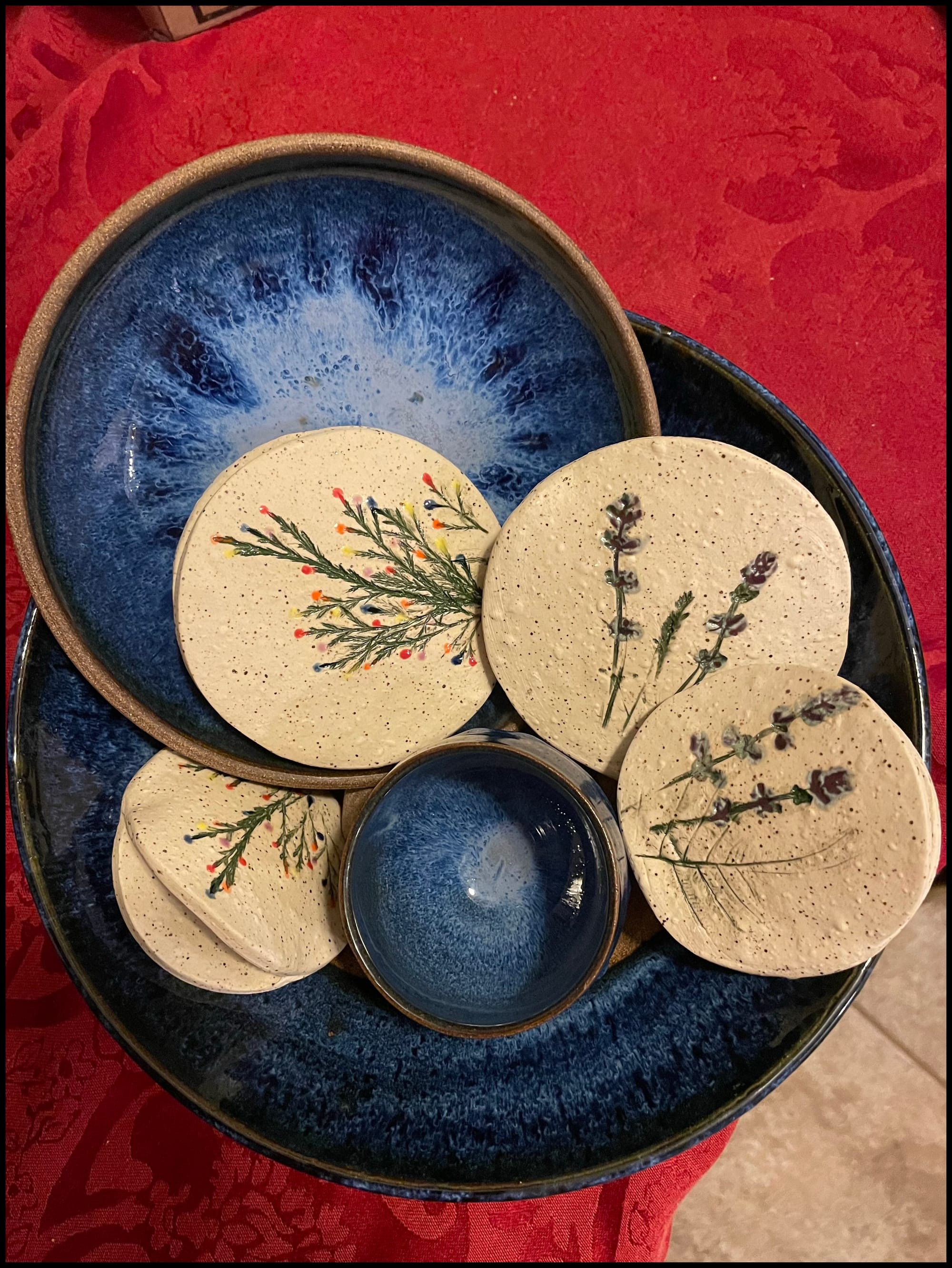Stacked blue-glazed ceramic bowls holding speckled cream dishes pressed with lavender and herb impressions, on a red tablecloth.