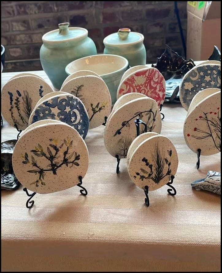 Round speckled ceramic coasters on black wire stands, pressed with plant impressions and star-and-moon patterns, beside pale green lidded jars.