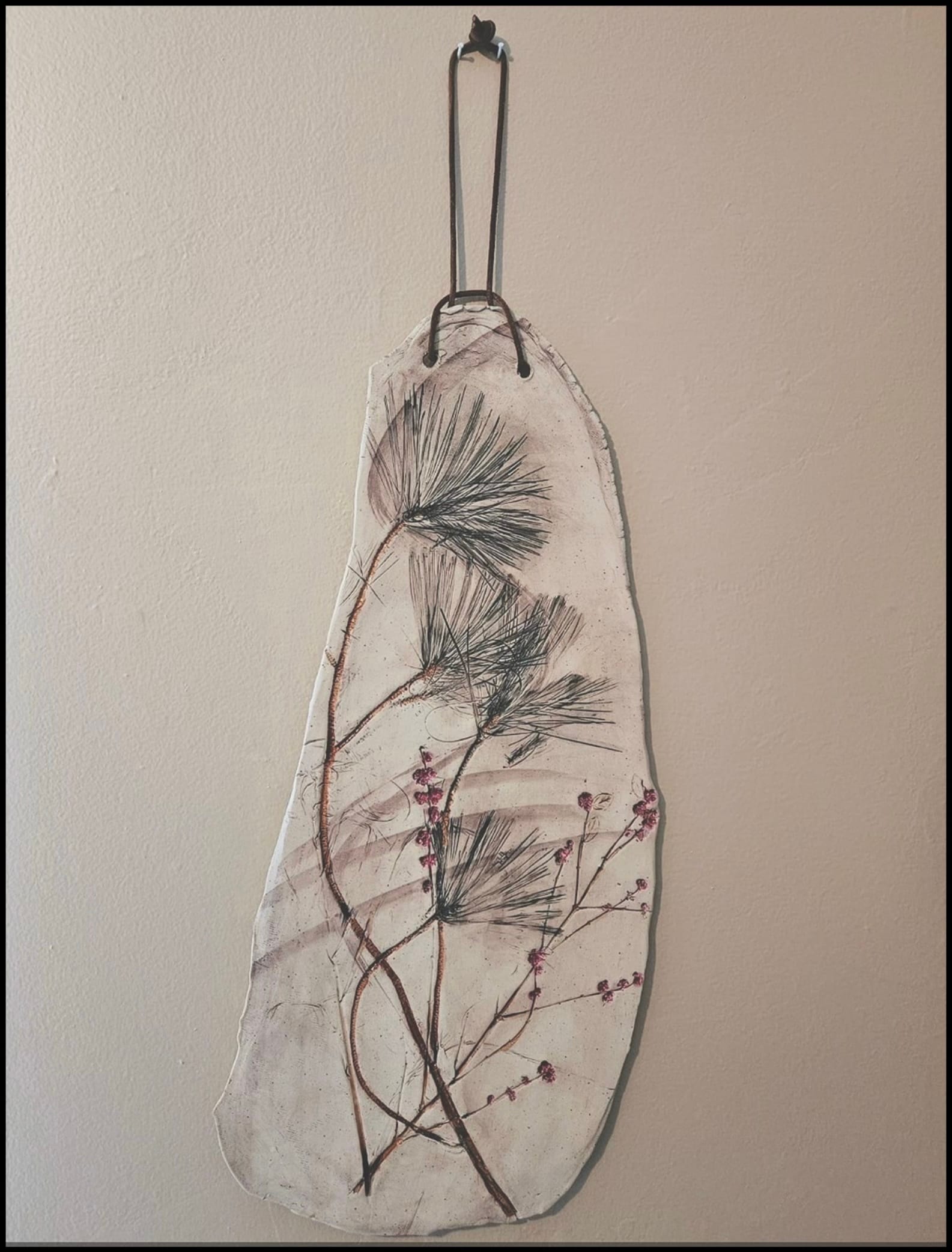 Wall-hung ceramic slab pressed with pine needle sprigs and pink berry branches, suspended by a leather cord from a hook.