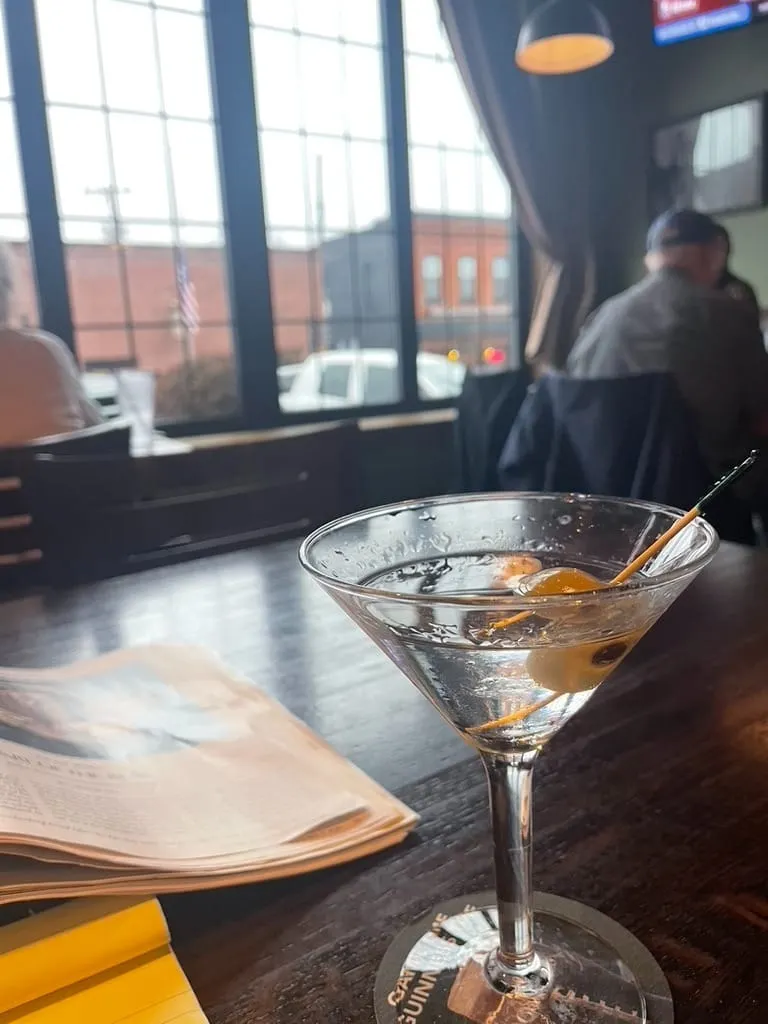 Close-up of a martini with an olive on a wooden table. The drink is backlit by the open front windows of the bar.