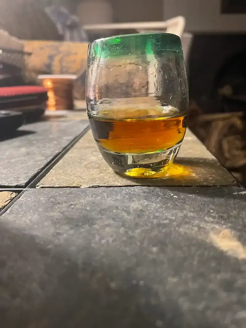 A glass filled with amber liquid sits on a tiled surface, casting a shadow. The glass's rim is green, and the setting is a cozy, dimly lit room.