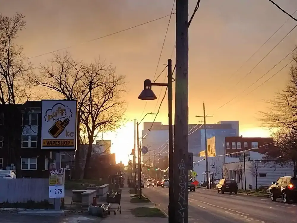 Urban street at sunset with warm glowing sky. Lamp posts and power lines frame the scene. A vape shop sign and bare trees line the sidewalk.