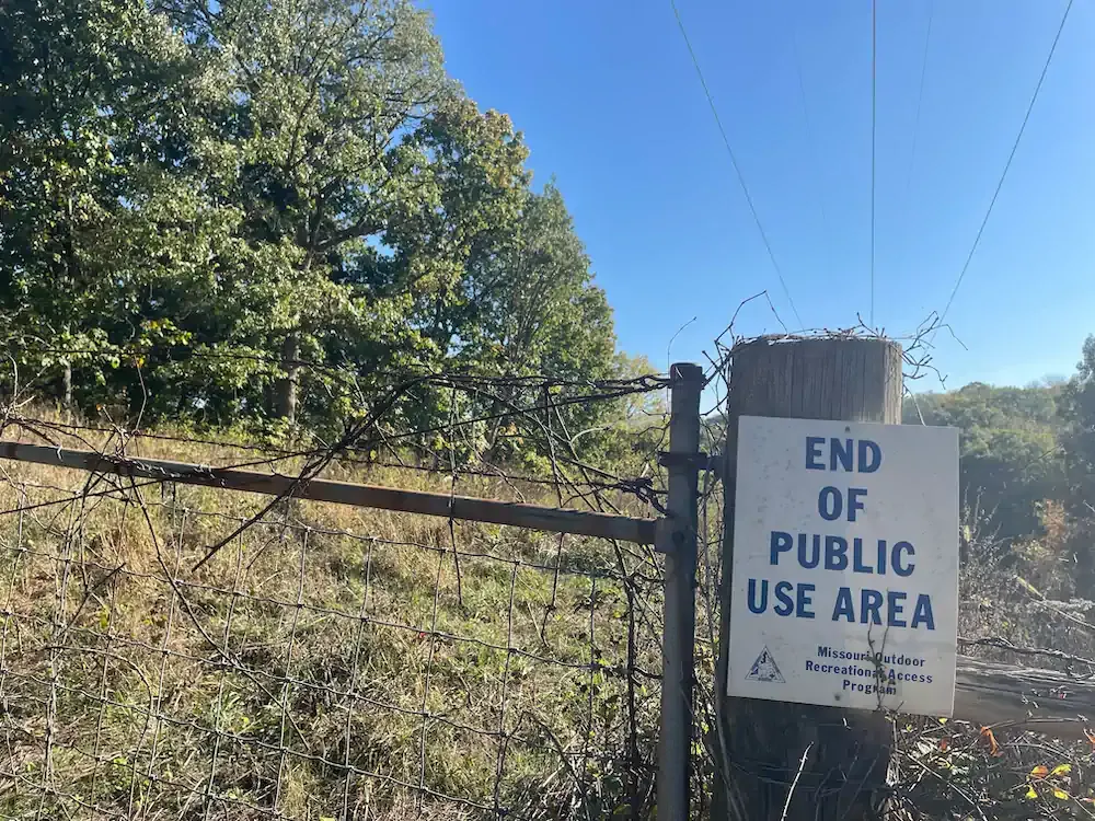 A barbed wire fence with a sign reading "End of Public Use Area" stands in front of a field and dense, sunlit trees under a clear blue sky.