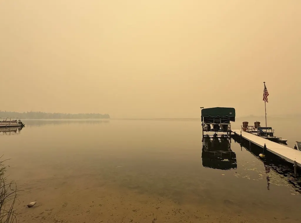 Smoky, hazy shoreline with a lakeside dock, covered boat lift, and American flag, sun reduced to a dim orange glow through wildfire smoke.