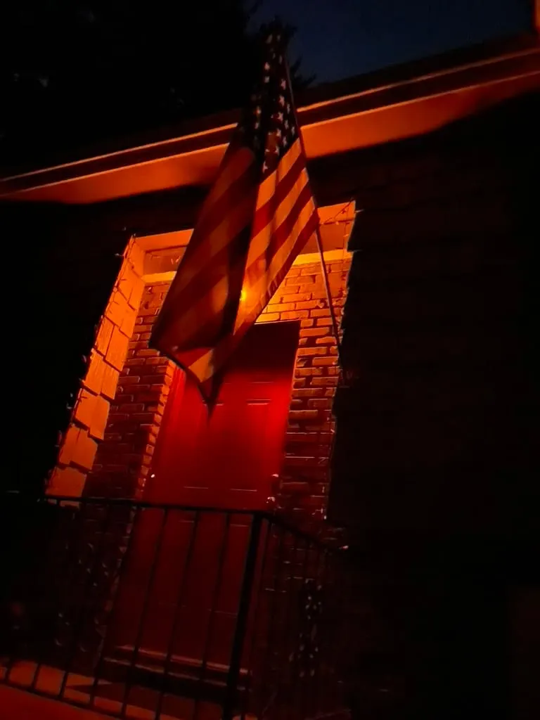 Photograph of an American flag hanging beside a red door on a brick house, illuminated warmly at night.