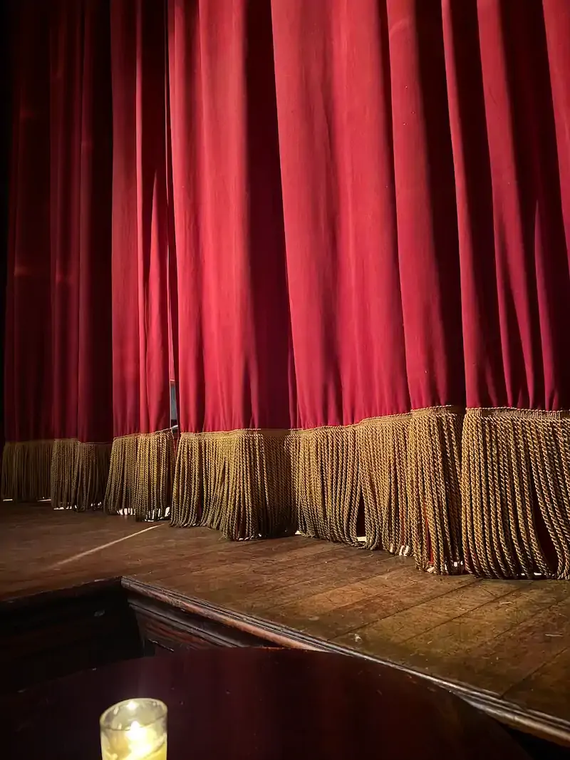 Deep red velvet stage curtains with gold fringe trim, viewed from the wings above a worn wooden stage and a lit candle.