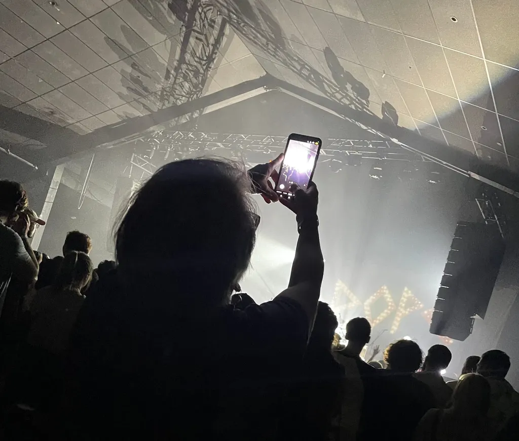Silhouetted concertgoer holds up a phone to film a hazy, smoke-filled venue, stage lights glowing behind illuminated letters spelling "POP."