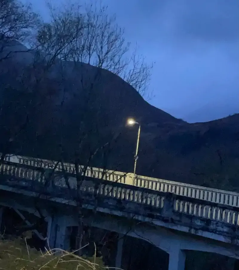 Dusk photograph of a white-railed concrete bridge lit by a single streetlamp, with bare trees and a dark mountain against a deep blue sky.