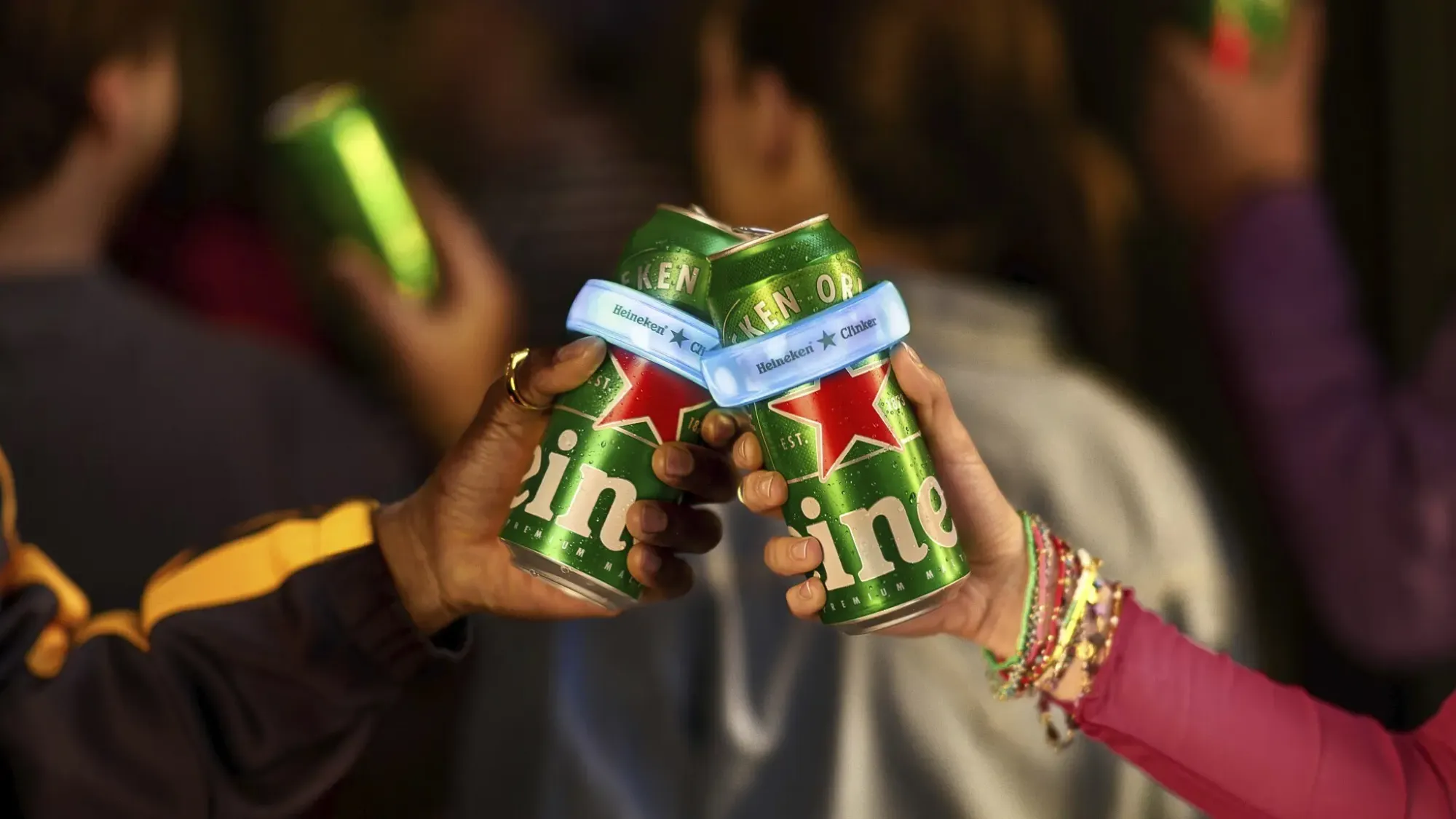 Deux festivaliers qui trinquent avec des canettes Heineken, le dispositif The Clinker accroché autour des canettes, ambiance Coachella en arrière-plan