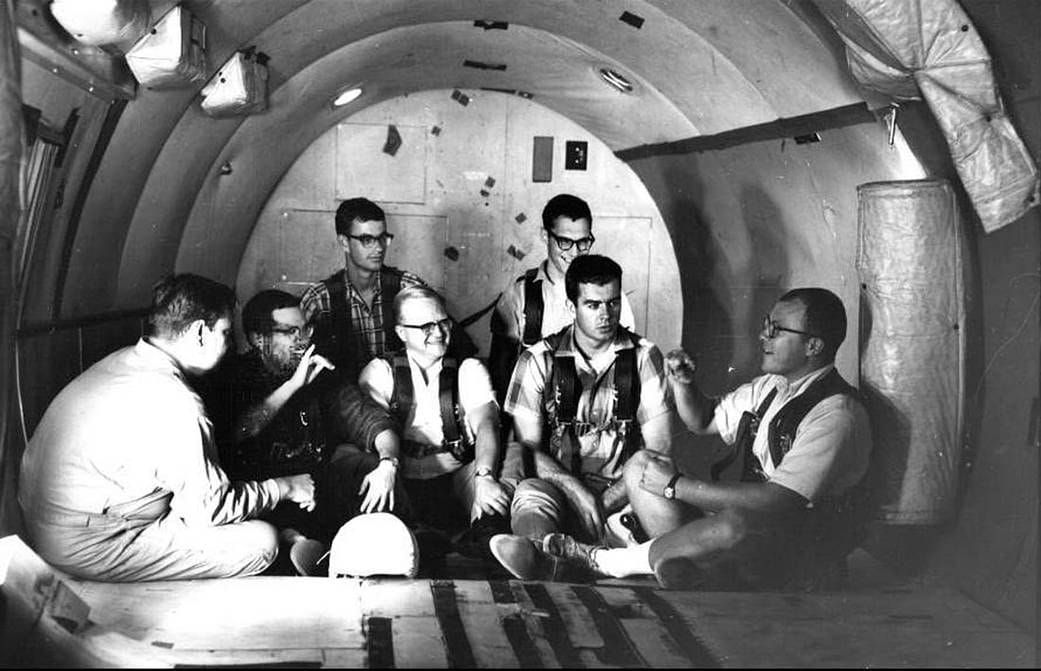 The photo is black-and-white and shows seven white men sitting on the floor of a cylindrical room, smiling and signing to each other. 