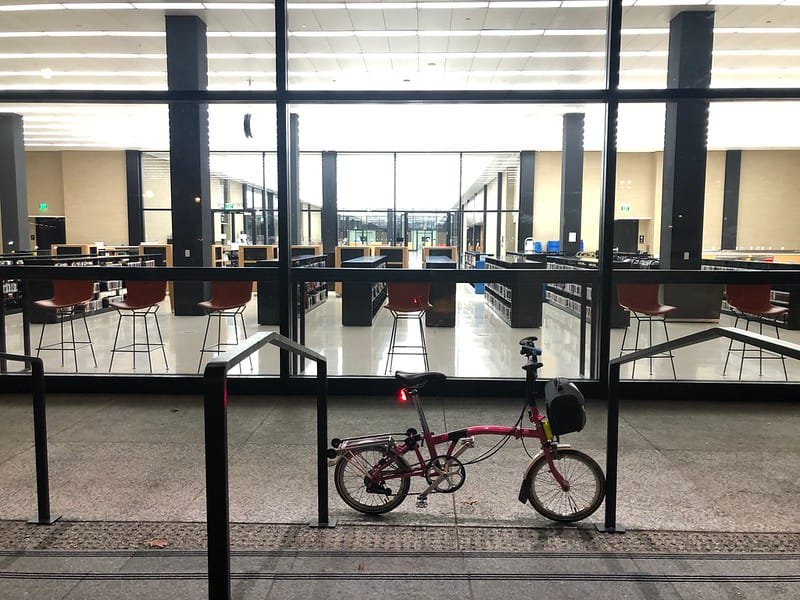 Exterior of MLK Library. A small red bike sits outside. You can see bookshelves and chairs through the glass wall.