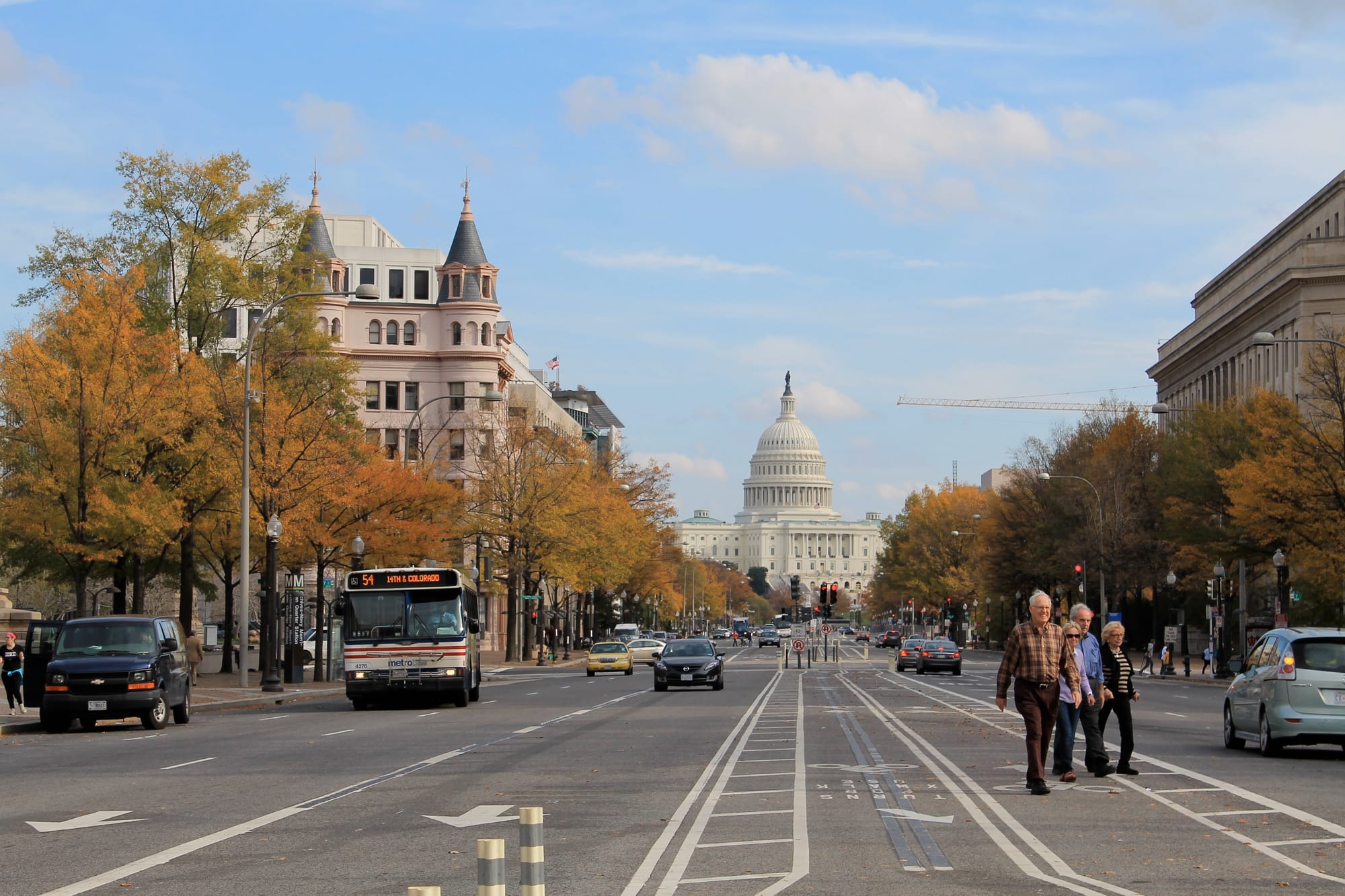 Four pedestrians cross the street with the U.S. Capitol building and fall foliage in the background.