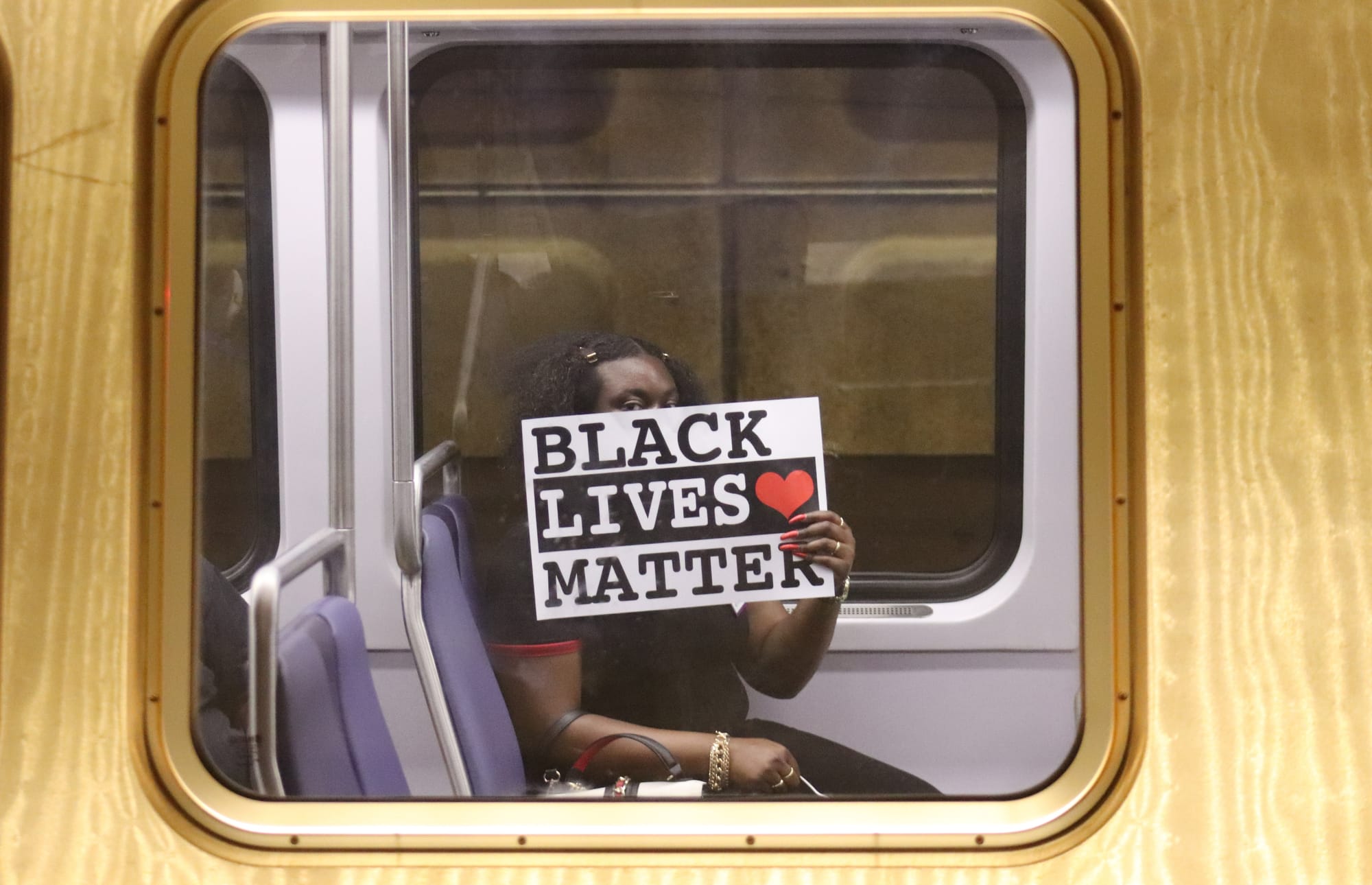 A black woman on a train holds a sign that reads "BLACK LIVES MATTER."