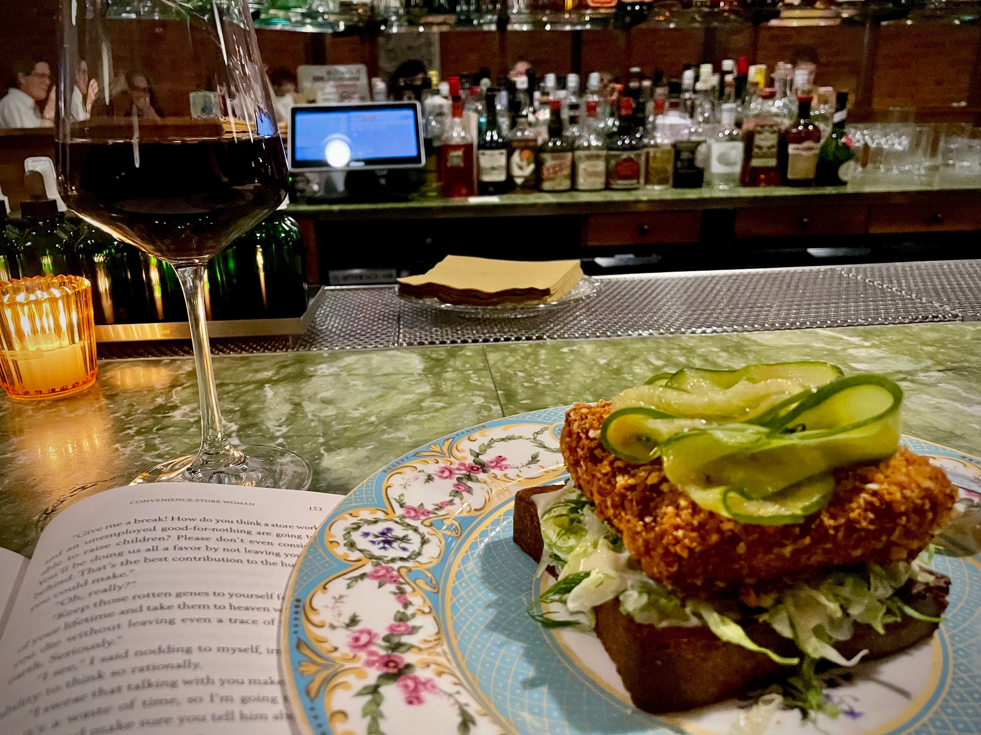 A glass of red wine, a book, and a sandwich on a bar counter. 