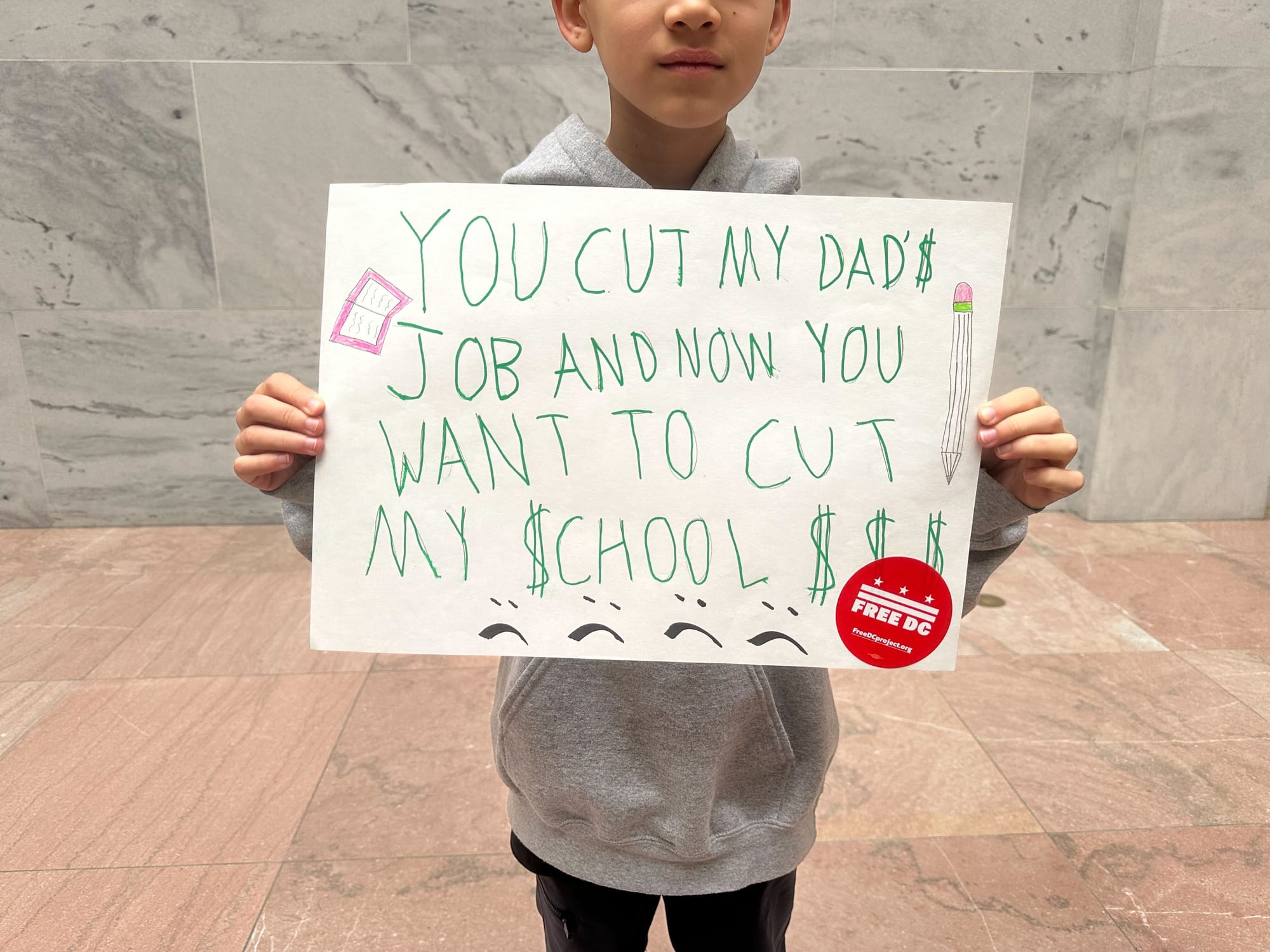 A picture of a kid holding a sign that says: "You cut my dad's job and now you want to cut my school $$$"