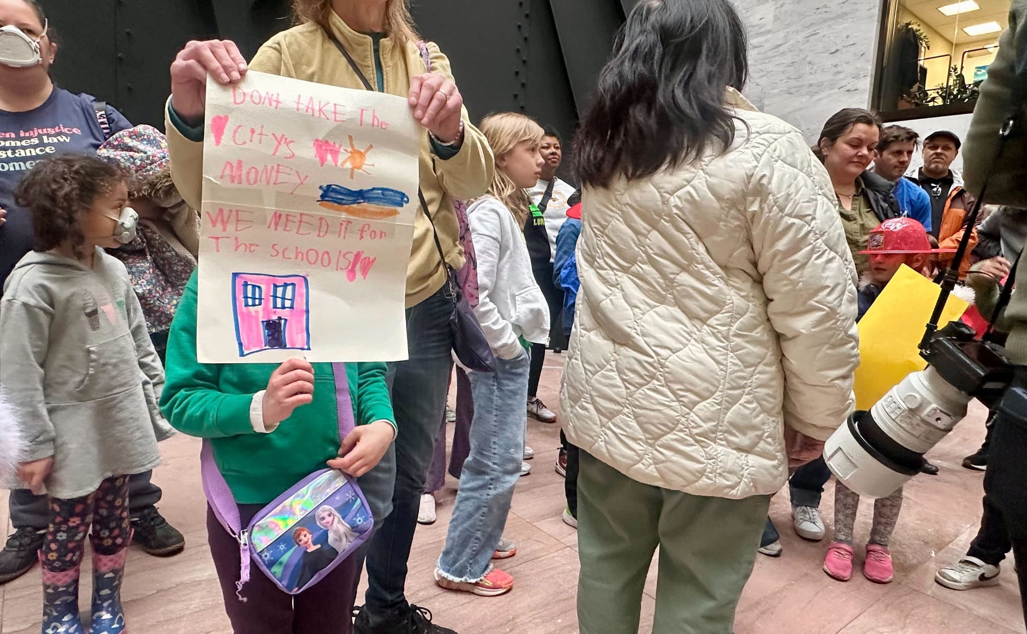 Kids and parents gather in the Senate, one holding a sign that reads: "Don't take the city's money we need it for the schools."