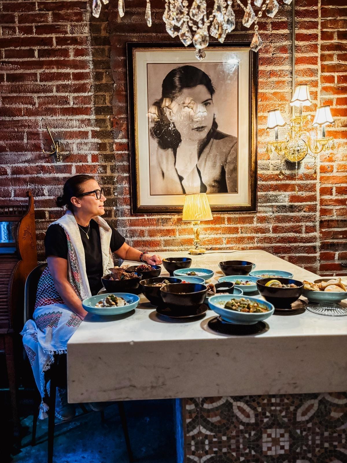 The owner of Nabiha, Nesrin Abaza, sits at a table covered in small plates of Palestinian food, with a black and white photograph of her mother behind her.