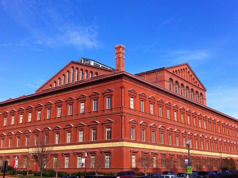 A phot of the The National Building Museum, a large, red brick building with a lot of windows, with blue skies in the background.