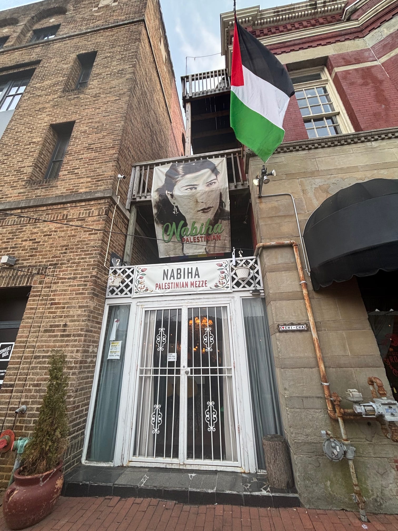  A large Palestinian flag hangs above the white and glass doors of a restaurant. A sign above the door says: "Nabiha Palestinian Mezze."