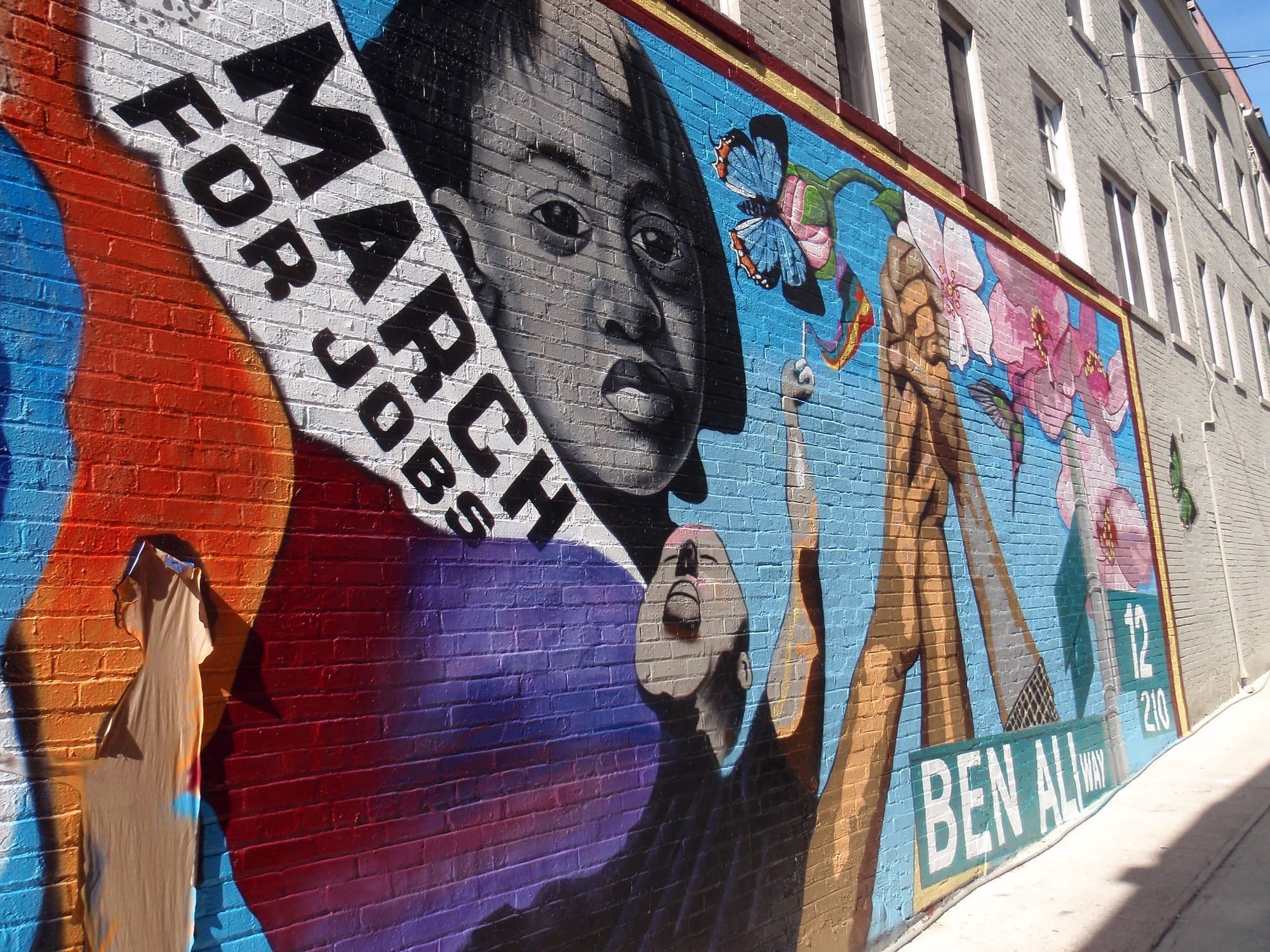 A mural of a Black woman next to a banner that reads March for Jobs, with a street sign that says Ben Ali Way. 