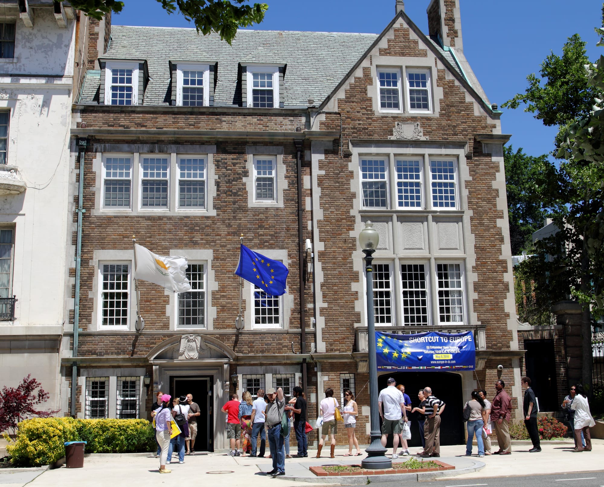 A small crowd is gathered outside The Embassy of the Republic of Cyprus, which is a narrow brown brick building.