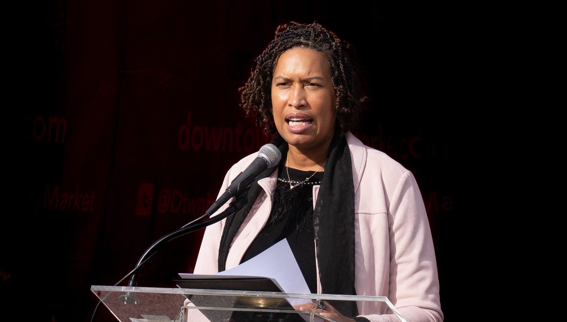 Mayor Muriel Bowser wearing a pink coat, standing before a podium speaking.