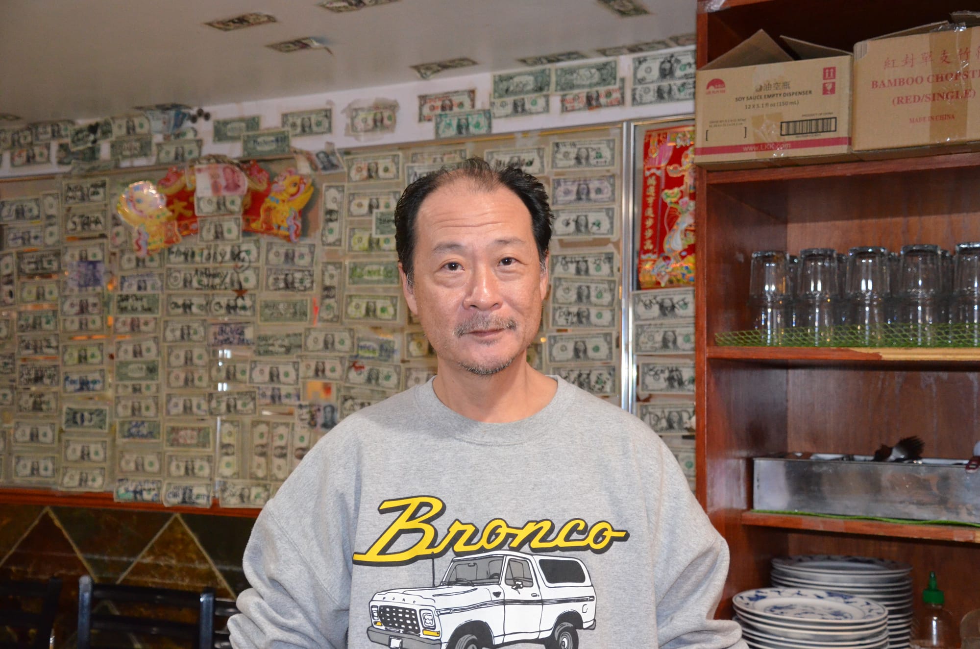 Peter Ko, owner of Jackey Cafe, poses for a photo. He's wearing a sweatshirt with "Bronco" written on it and stands in front of a money wall 