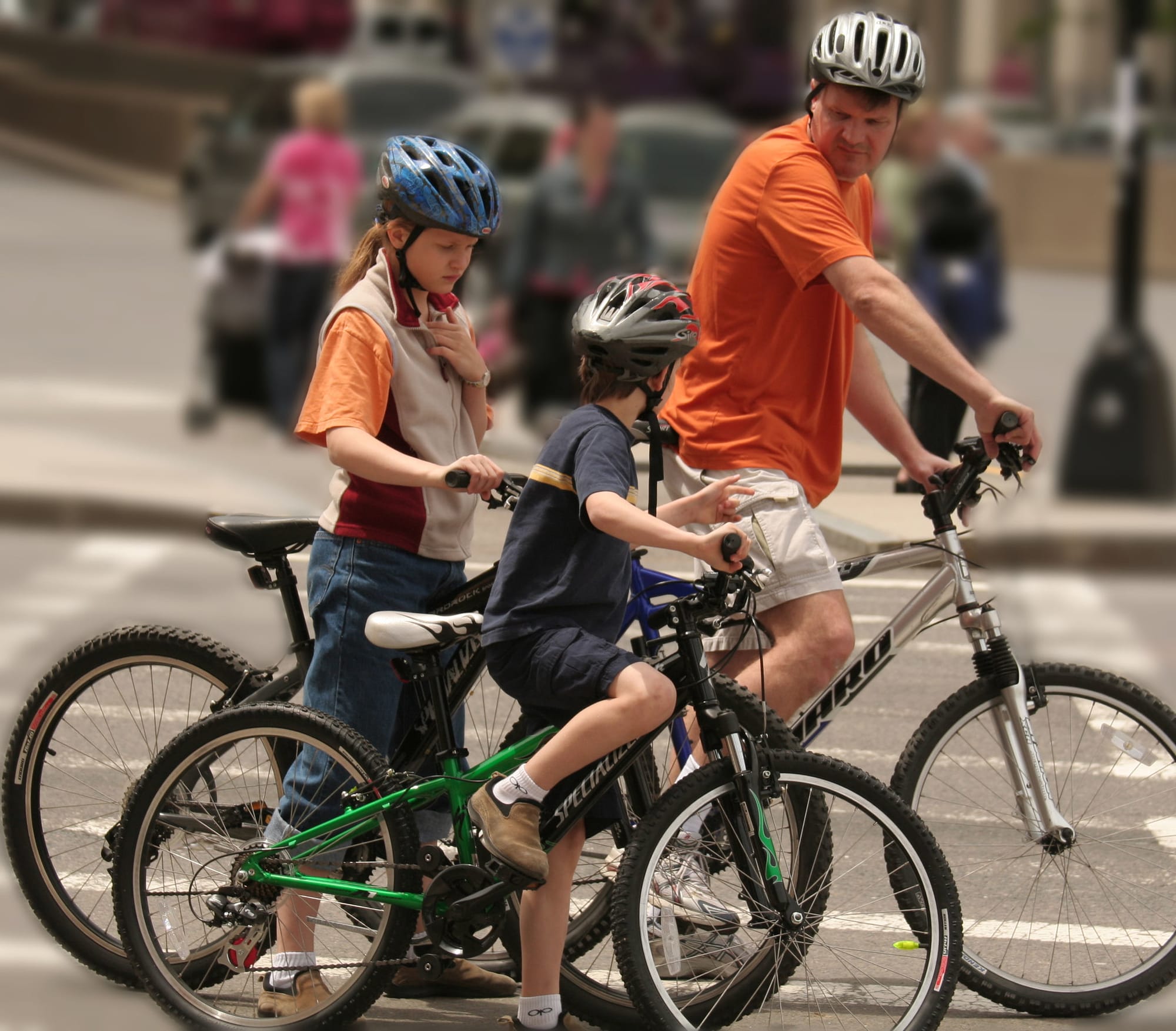A young boy, young girl, and adult man riding bikes, paused in a crosswalk. 