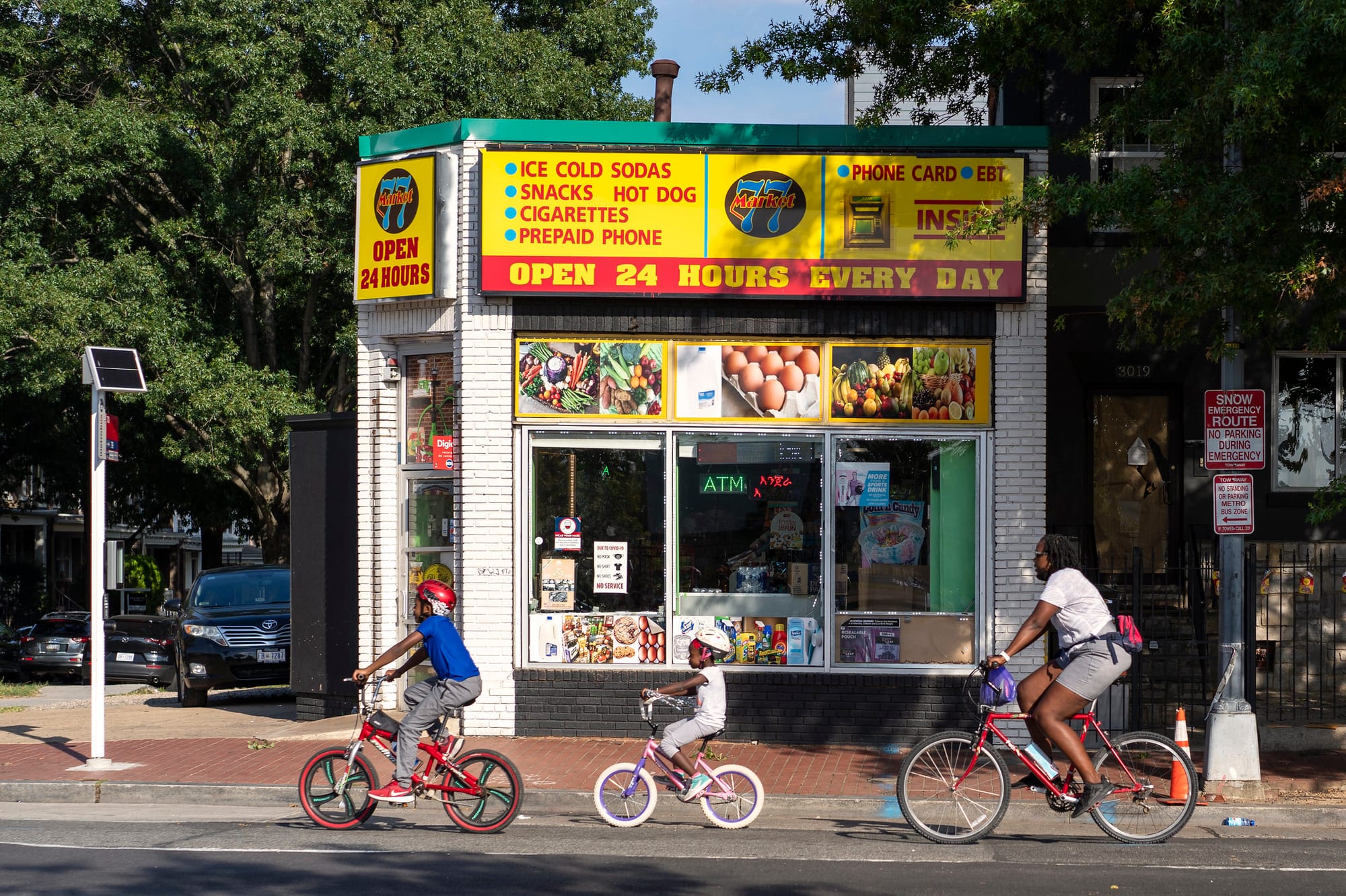 A young boy, girl, and adult woman riding bikes in a row past a corner store. 