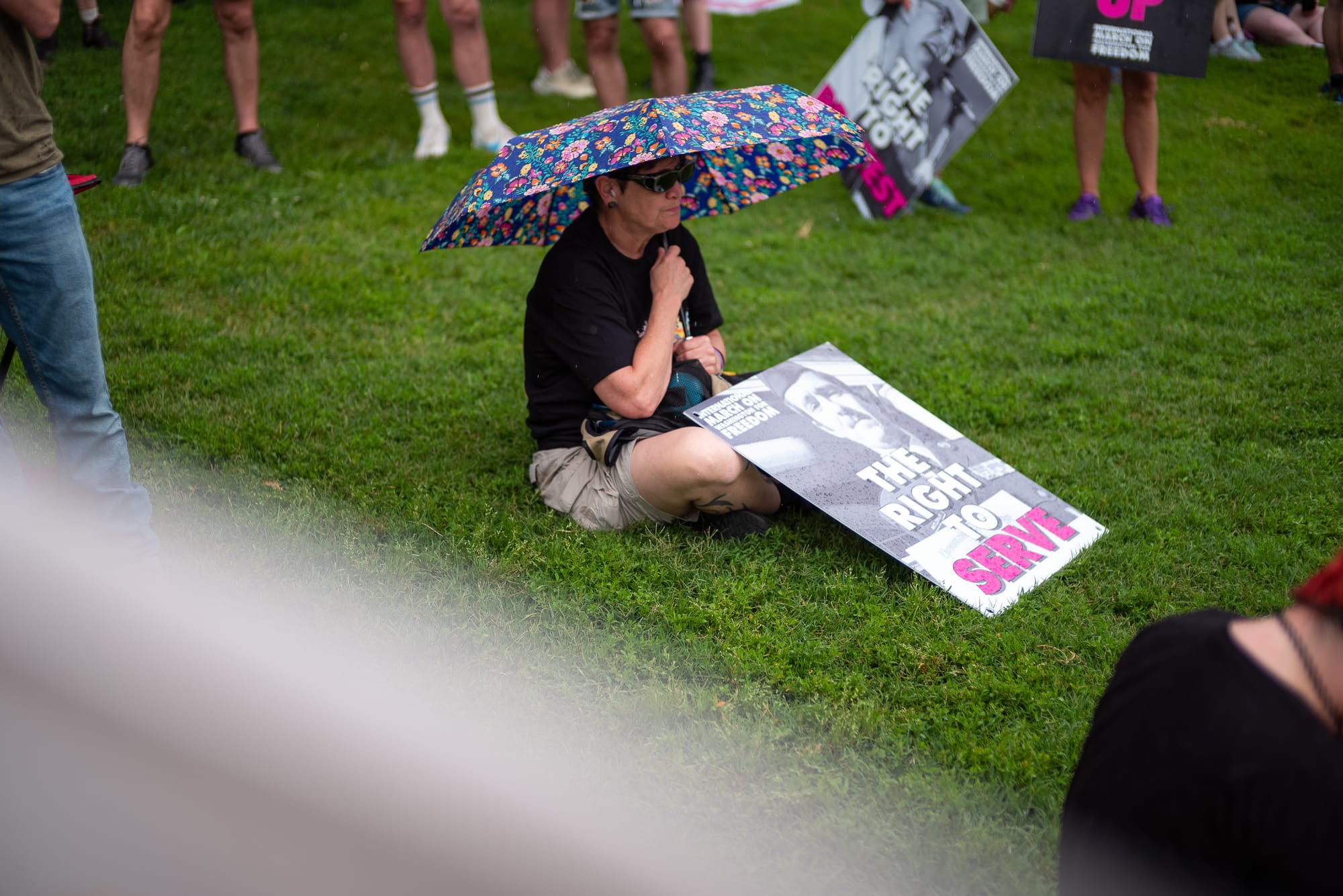 A person sitting holding an umbrella in the grass, with a sign that reads The Right to Serve.