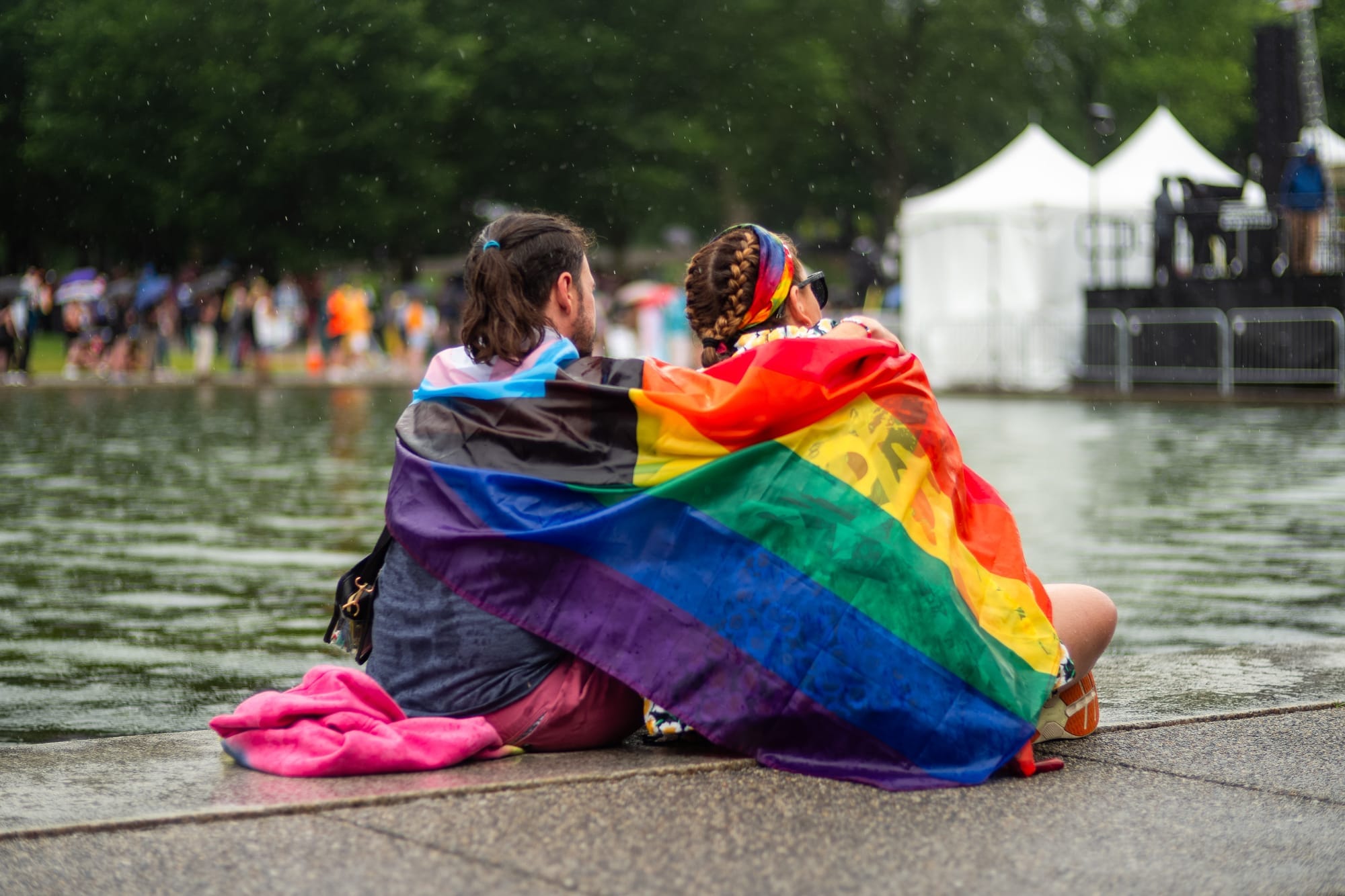 Two people draped in an LGBTQ flag/blanket along a water feature on the National Mall. 
