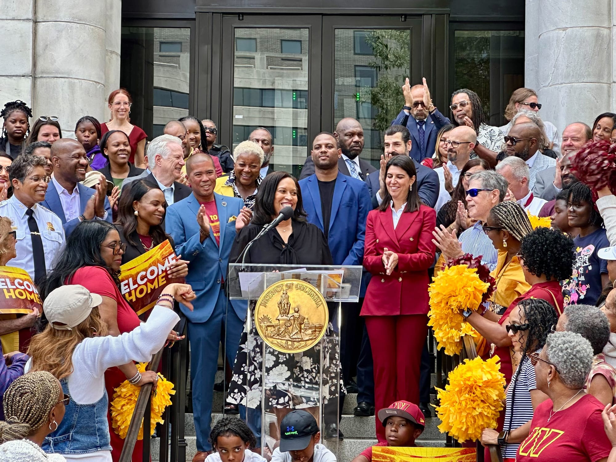 Mayor Bowser speaking before a podium, surrounded by people in Commanders gear and colors. 