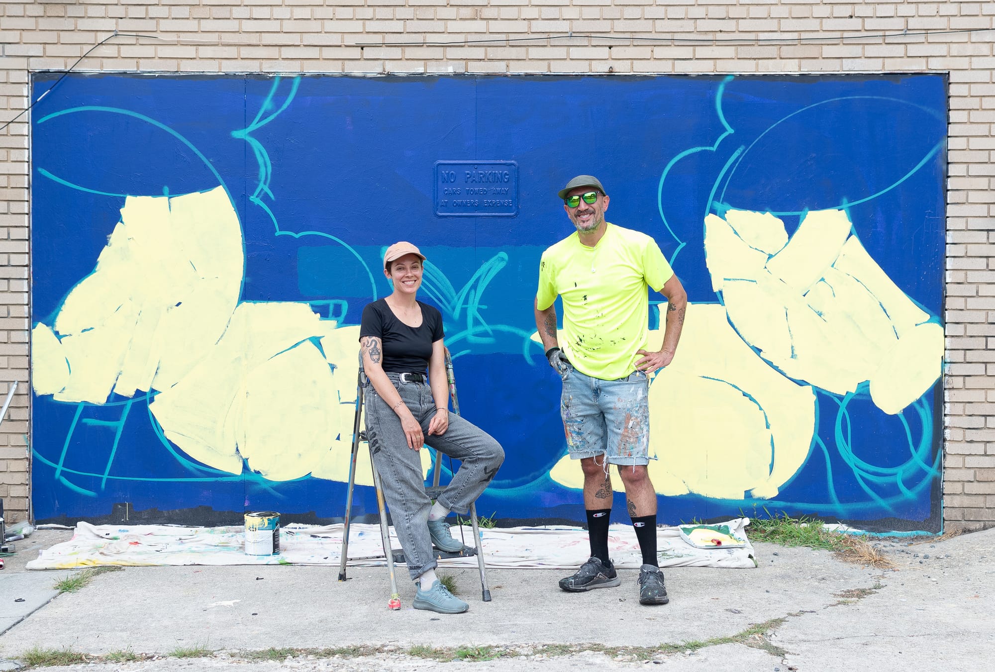 Two people in painting clothes standing in front of a blue wall as its getting painted. 
