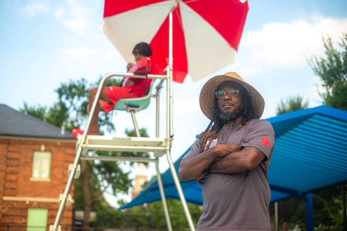 Man wearing hat with arms folded standing next to lifeguard stand at pool.