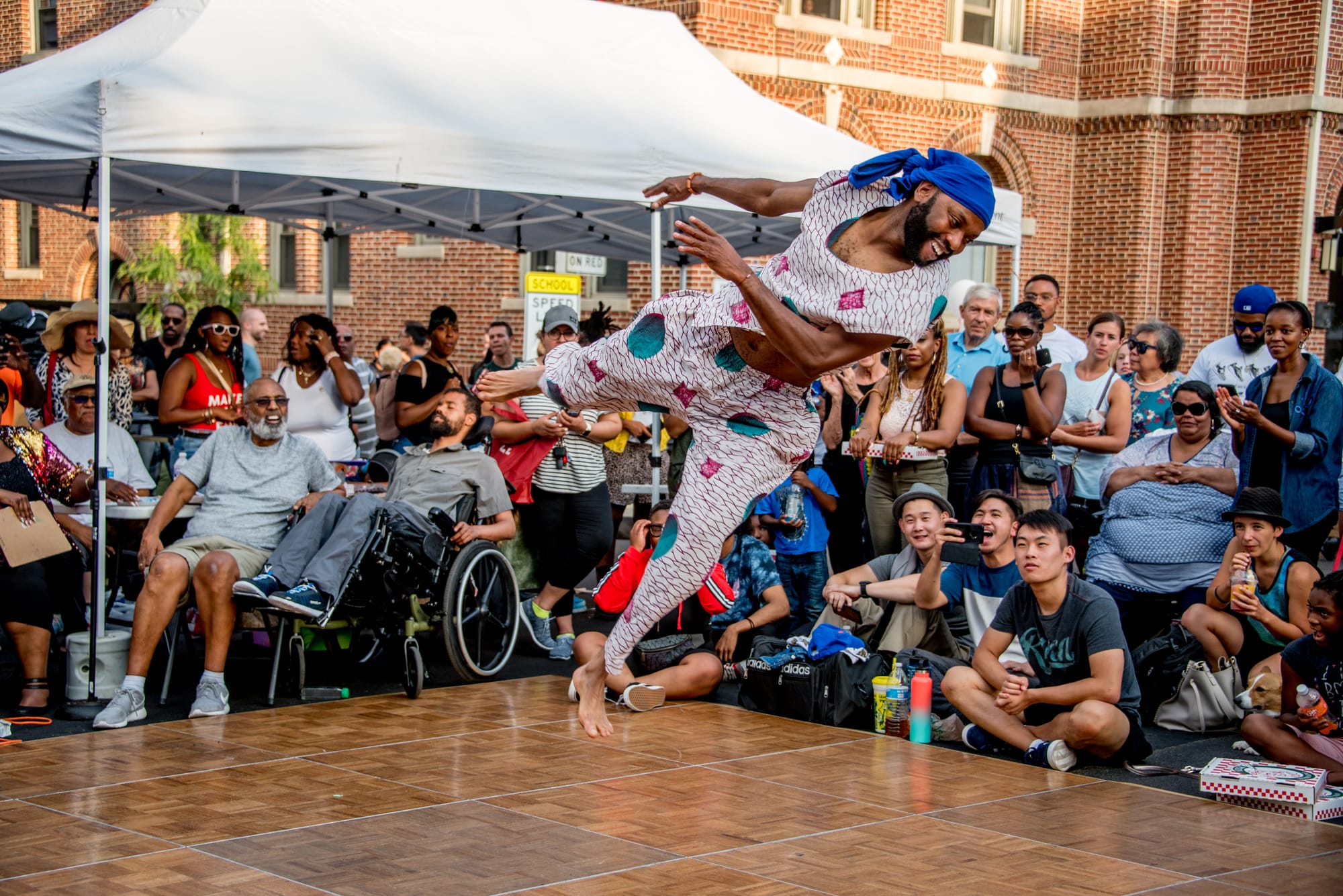 Someone wearing African print dances on a wooden stage in midair, with a large crowd of excited onlookers.