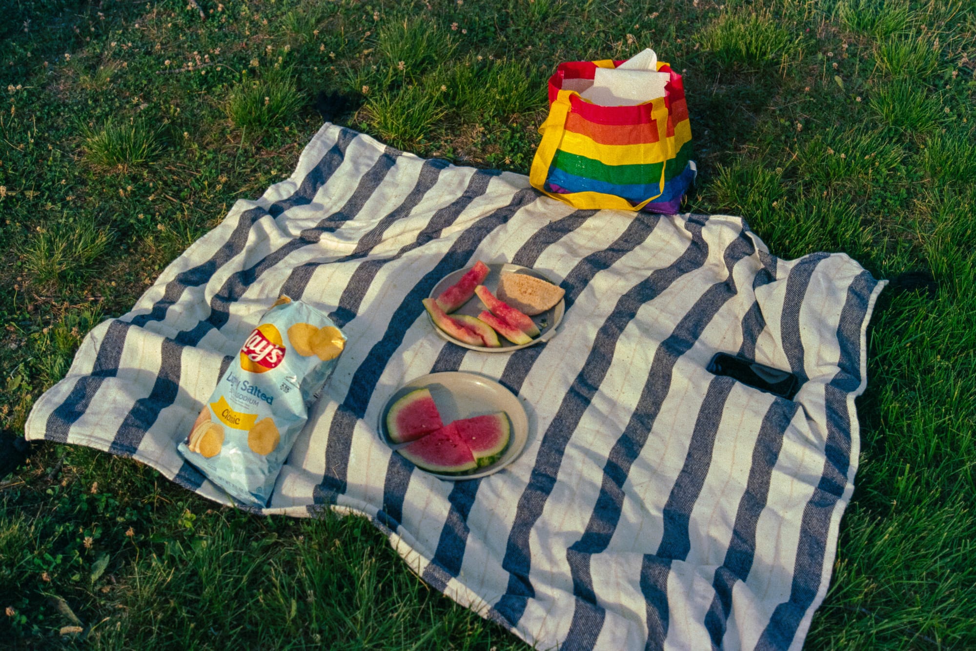 A striped picnic blanket on green grass with a rainbow tote bag, a plate of watermelon, and a bag of Lay's Potato chips.