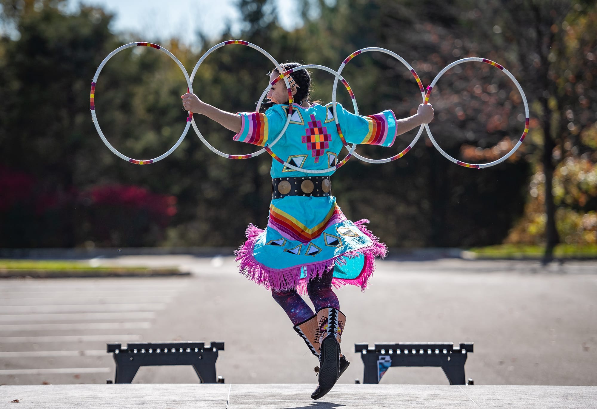 A hoop dancer performs, wearing traditional clothing and holding five hoops on their arms and shoulders.